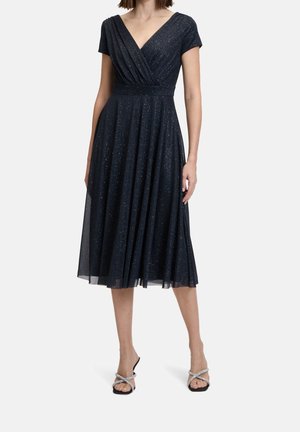 Kobieta w granatowej, błyszczącej sukience midi bez rękawów z dekoltem w kształcie litery V i plisowaną spódnicą, w połączeniu z czarnymi sandałami na paski.