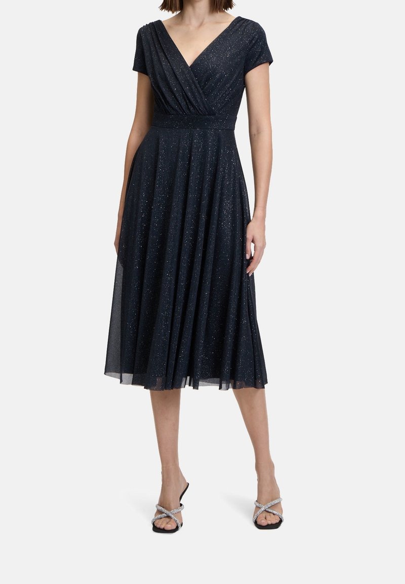 Femme portant une robe midi sans manches bleu marine pailletée avec un décolleté en V et une jupe plissée, associée à des talons noirs à lanières.