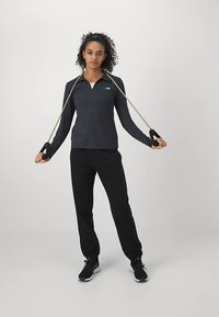 Top deportivo de manga larga en gris oscuro con cuello, combinado con pantalones negros. La modelo sostiene bandas de ejercicio con mangos negros y lleva zapatillas deportivas negras.