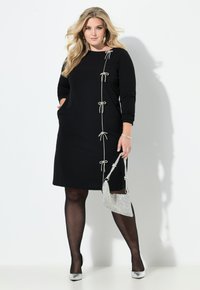Vestido negro de manga larga, con lazo decorativos a lo largo de un lado. Combinado con un bolso plateado y zapatos metálicos. Textura de tela suave.