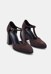 Zapatos de tacón alto de ante marrón con diseño de tira en T y hebilla, que presentan un tacón en bloque texturizado. Forma clásica de punta afilada.