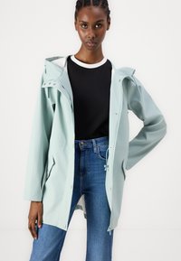 Lys blå vanntett jakke med hette, trykknapplukking og snor i livet, kombinert med en svart topp og blå jeans.