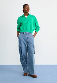 Sweatshirt verde com fecho éclair e gola alta, acompanhado de calças cargo azuis claras com bolsos laterais e corte largo. Sapatos castanhos.