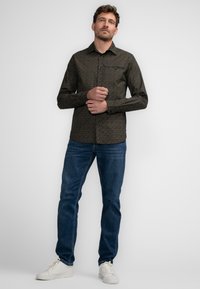 Langarmig, donker patroon shirt met een button-down kraag, gecombineerd met donkerblauwe jeans en witte sneakers, staand tegen een lichte achtergrond.