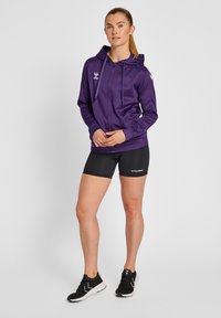 Felpa con zip viola dotata di cappuccio, con dettagli neri e logo, abbinata a pantaloni sportivi neri e sneakers nere. Texture del tessuto liscia.