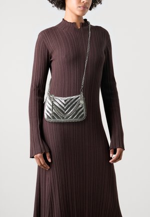 Mujer con vestido marrón de manga larga de canalé, llevando un pequeño bolso cruzado plateado acolchado con correas de cadena.