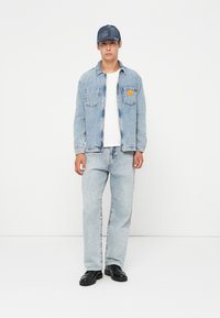 Light blue denim jacket with front pockets, paired with matching light blue jeans. White T-shirt underneath, black chunky shoes, and a denim cap.