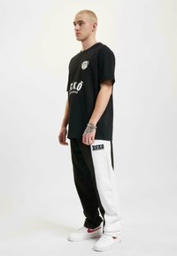 Black T-shirt with logo and text, paired with black and white sweatpants, featuring side stripes and branding. White sneakers complete the look.