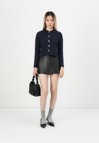 Marineblauwe cropped cardigan met een kraag en gouden knopen, gecombineerd met een zwarte leren minirok. Zwarte tas met een gestructureerde afwerking en grijze sokken.