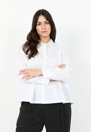 Žena s dlouhými tmavými vlasy, oblečená v bílé košili na knoflíky a černé kalhoty s proužky, stojící se založenýma rukama proti bílému pozadí.