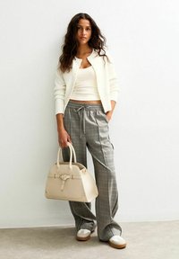 Vit cardigan över en figurnära vit linne, i kombination med grå rutiga vida byxor. Bär en ljusbeige handväska med strukturerad design.