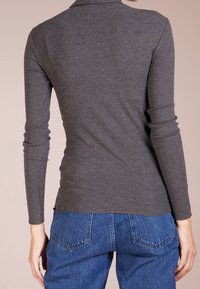 Eine Frau trägt ein enganliegendes graues Langarmoberteil und blaue Jeans, von hinten gegen einen neutralen Hintergrund gezeigt.