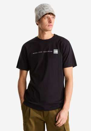 Joven hombre con un gorro beige y una camiseta negra con el texto "Never Stop Exploring", de pie con las manos en los bolsillos sobre un fondo blanco.