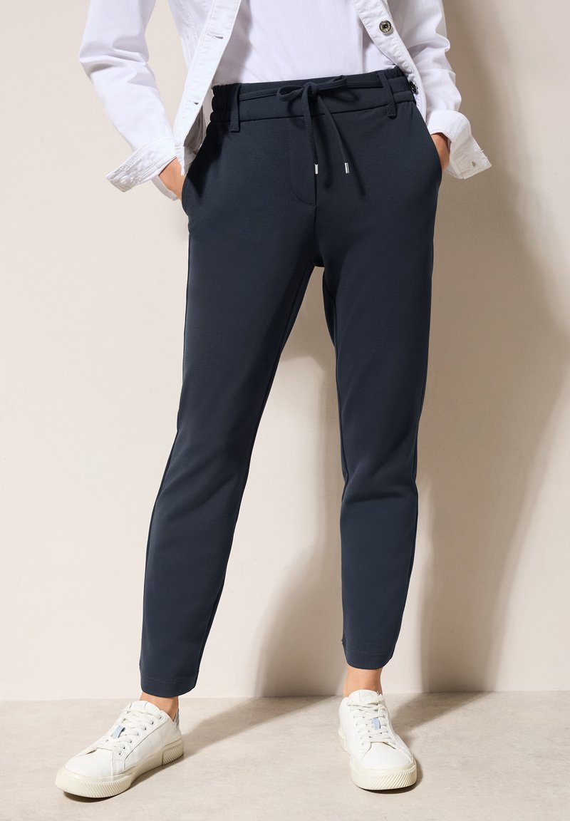 Pantalons de jogging bleu marine avec une taille élastique et un cordon de serrage, dotés de jambes fuselées et de poches latérales, associés à des baskets blanches.