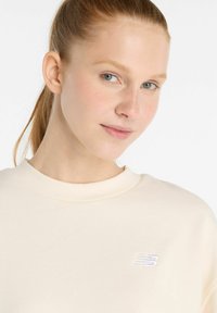 Sudadera color crema con cuello de canalé y un pequeño logo blanco en el pecho. Textura suave, corte casual, diseño minimalista.