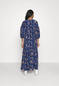 Robe maxi bleue à fleurs avec des manches bouffantes, cintrée à la taille, présentant une jupe à volants et un tissu doux et léger. Baskets blanches visibles.