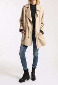Trench beige con un cuello ancho, cintura elástica y dos bolsillos laterales. Llevado sobre un suéter oscuro, combinado con jeans azules rasgados y botas negras.