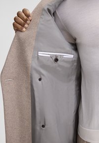 Blazer in lana beige con foderatura grigia liscia, dotato di due bottoni marroni e una tasca sul petto con bordo bianco. Tessuto esterno texture.