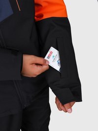 Mani che sistemano un pass per sci con loghi in una tasca con zip sulla manica di un giubbotto invernale nero e arancione.