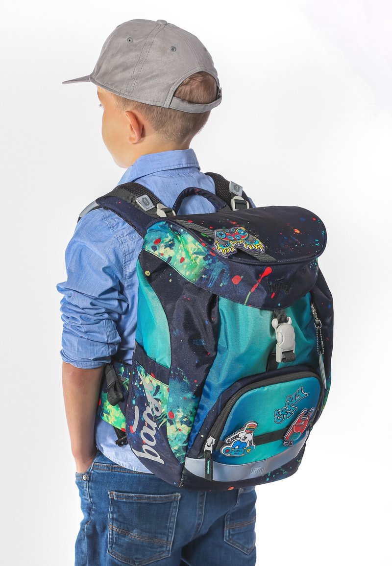 Kind met een grijze pet en een blauw shirt draagt een kleurrijke rugzak met een gespat verfdesign en verschillende patches.