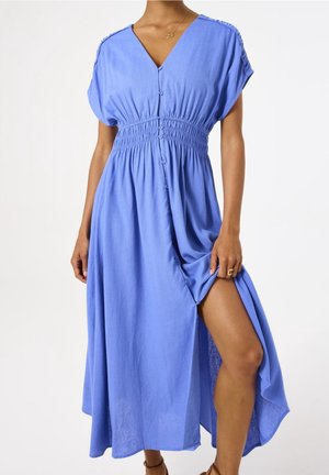 Femme portant une robe midi bleue boutonnée avec manches courtes et taille cintrée, soulevant la jupe pour dévoiler sa jambe.