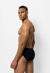Zwarte zwemslips van opzij getoond, met een gladde textuur en een elastische tailleband. Het model heeft een gespierd postuur en kort haar.