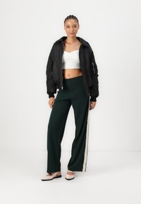 Zwarte bomberjack, witte cropped top, groene wijde broek met crèmekleurige zijsstripes en beige schoenen met riempjes. Soepele texturen en ontspannen pasvorm.