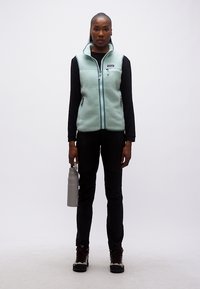 Γυναίκα που φοράει ανοιχτό μπλε γιλέκο από φλις, μαύρο μακρυμάνικο πουκάμισο, μαύρο παντελόνι, παπούτσια πεζοπορίας, κρατώντας γκρι παγούρι, στέκεται μπροστά σε λευκό φόντο.