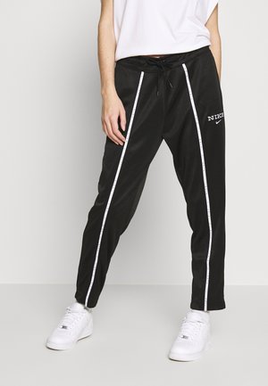 Persoon draagt zwarte Nike trainingsbroek met witte zijstrepen en witte sneakers, staand tegen een effen lichte achtergrond.