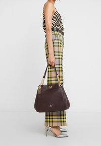 Brązowa skórzana torebka z łańcuszkowymi ozdobami, o zaokrąglonym kształcie i złot�ą plakietką z logo. Torebka jest trzymana przez osobę noszącą kratkowane spodnie.