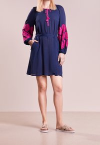 Marinblå klänning med långa rosa broderade ärmar, dragsko i halsen och midjeband. Inkluderar sidofickor. Bärs med lätta, glittrande sandaler.