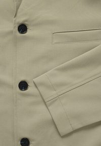 Világos olívazöld dzseki sima textúrával, fekete gombokkal, mellzsebbel és szabott ujjal, varrott díszítésekkel.