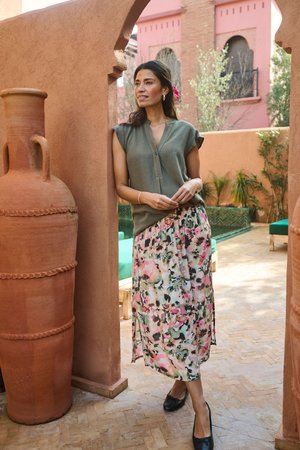 Femme debout dans une cour ensoleillée avec des murs en terre cuite, portant un haut vert sans manches, une jupe fleurie, des chaussures noires, avec une fleur rose dans les cheveux.