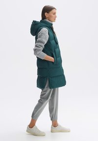 Gestepptes petrolfarbenes Vest mit einer passgenauen Kapuze, vorderen Taschen und einem Reißverschluss, kombiniert mit einem hellgrauen Strickpullover und grauen, schmal zulaufenden Hosen.