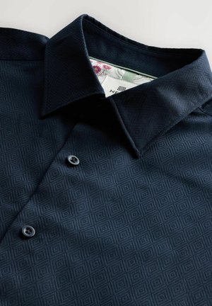 Camicia abbottonata blu navy con un motivo geometrico, tessuto testurizzato e colletto ripiegato con fodera interna a contrasto. Due bottoni neri visibili.