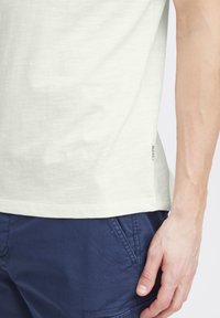 Camisa de color claro y de manga corta, hecha de una tela suave, con una etiqueta lateral y un dobladillo ligeramente curvado, combinada con pantalones cortos color marino.