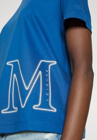 Camiseta azul de manga corta con una gran "M" blanca contornada y la palabra "LEISURE" impresa a un lado. Textura suave, diseño casual.
