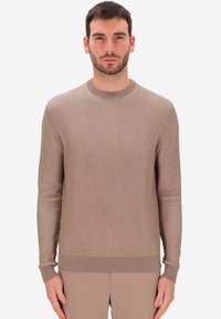 Uomo con capelli corti scuri e barba, che indossa un maglione a maniche lunghe ribattuto beige e pantaloni beige coordinati, in piedi contro uno sfondo bianco.