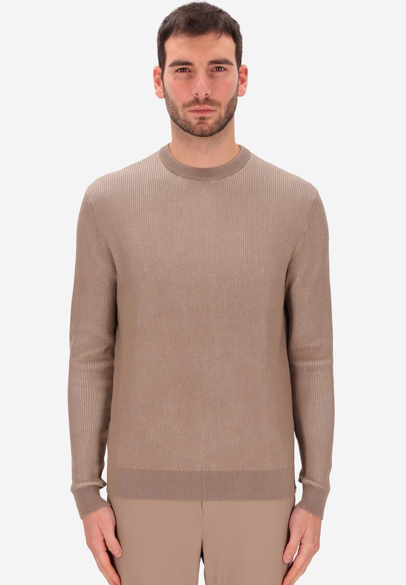 Uomo con capelli corti scuri e barba, che indossa un maglione a maniche lunghe ribattuto beige e pantaloni beige coordinati, in piedi contro uno sfondo bianco.