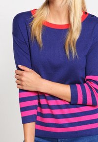 Marinblå och rosa randig tröja med trekvartslånga ärmar, har en rund halsringning med en röd accent och detalj med tre knappar på axeln.