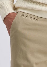 Pantalons en coton beige avec une texture lisse et un détail de poche inclinée, associés à un pull à manches côtelées crème.