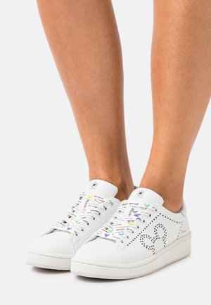 Witte sneakers met geperforeerd ontwerp van een muizenhoofd aan de zijkanten en bedrukte veters, gedragen op blote voeten, tegen een effen lichte achtergrond.