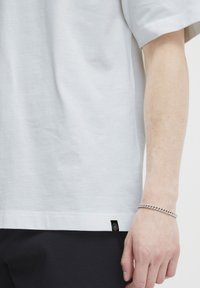 Bras droit d'une personne portant une chemise blanche à manches courtes simple avec une petite étiquette noire près de l'ourlet et un bracelet en chaîne argenté.