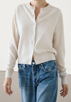 Personne portant un cardigan crème boutonné et un jean taille haute bleu, debout à l'intérieur avec les bras détendus le long du corps.