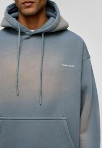 Grijsblauwe hoodie van zacht materiaal. Bevat een kangoeroezak, trekkoord en een klein "YOUNG ATELIER" logo op de borst.