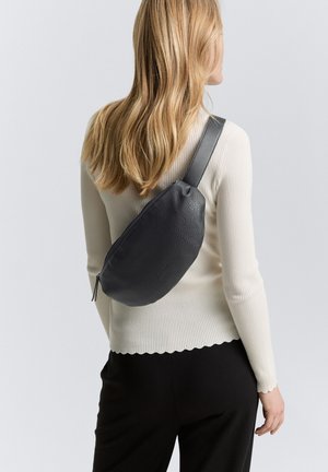 Sac en cuir noir à bandoulière avec une finition texturée, fermeture zippée et une fine sangle, porté en bandoulière sur un fond clair.