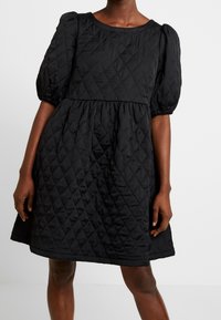 Schwarzes, gestepptes Kleid mit Puffärmeln, rundem Ausschnitt, gerafftem Bund und knielangem Saum. Das Material hat eine glatte, gepolsterte Textur.