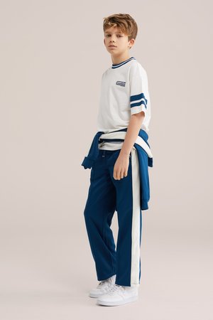 Fant, ki stoji, oblečen v belo majico s modrimi črtami, modre športne hlače z belo stransko črto, bele superge in zavezano modro jakno.