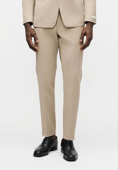 Άνδρας που φοράει μπεζ παντελόνι και σακάκι κοστουμιού, με μαύρα δερμάτινα παπούτσια, στέκεται μπροστά σε λευκό φόντο.