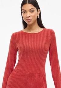 Červené žebrované šaty s dlouhým rukávem, kulatým výstřihem a přiléhavým tělem, přecházející do rozšířené sukně. Hladká textura s jemným leskem.